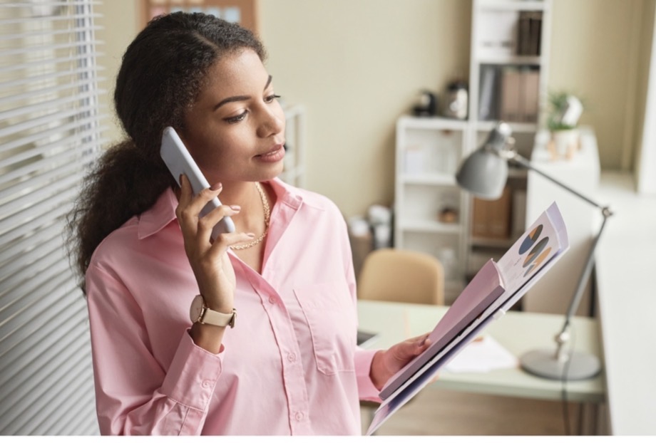 Young Black woman in a soft pink blouse reviewing a report while on a phone call in a light-filled office.