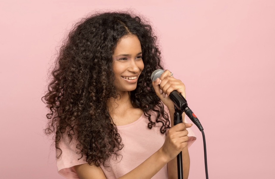 Young woman speaking into a microphone on a soft pink backdrop.